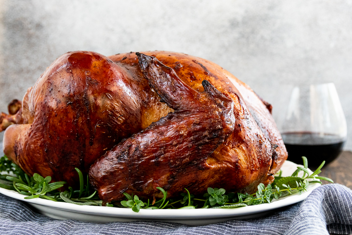 golden brown smoked thanksgiving turkey on a white platter surrounded by a fresh herb garnish