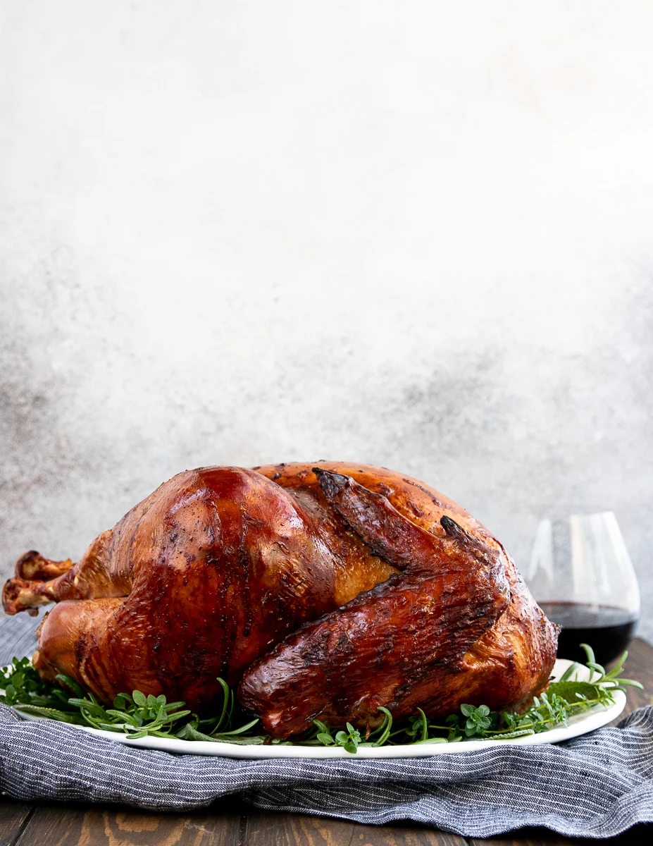 side view of a whole thanksgiving turkey on a white platter sitting on a grey pinstripe napkin with a glass of pinot noir in the background