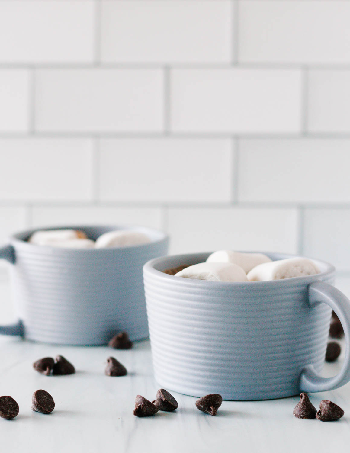 two grey textured mugs with hot chocolate on a light counter with a white subway tile background