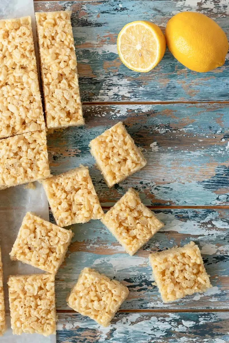flat lay photo of the lemon cereal treats on a blue mottled background with a sliced lemon in the upper right corner and scattered squares on the lower left