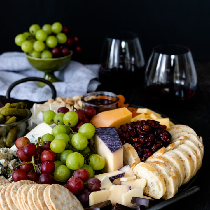 round cheese board with grapes, bread, and crackers. Wine glasses in the background