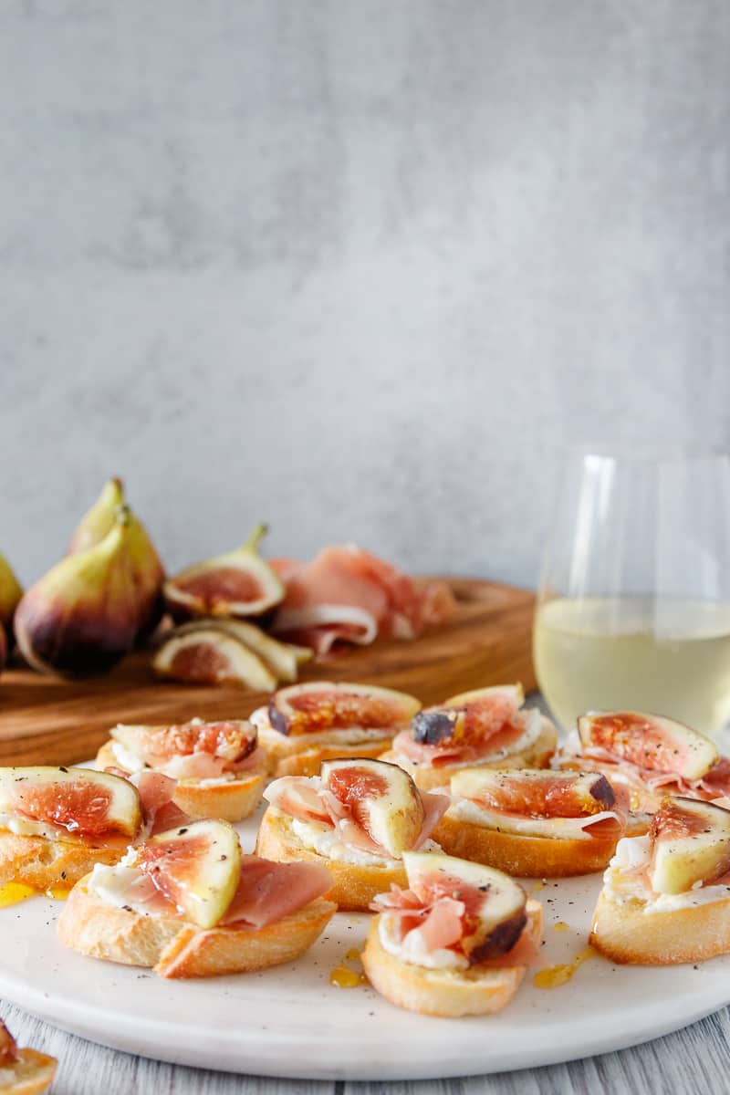 fig crostini on a marble tray with a glass of white wine and more fresh figs in the background