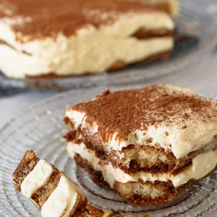 angled picture of a piece of tiramisu with a corner broken off and sitting on a fork at the edge of the plate