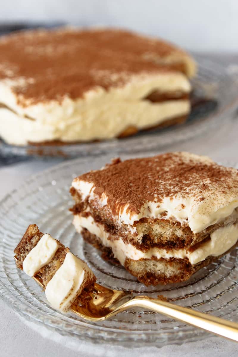 angled picture of a piece of tiramisu with a corner broken off and sitting on a fork at the edge of the plate