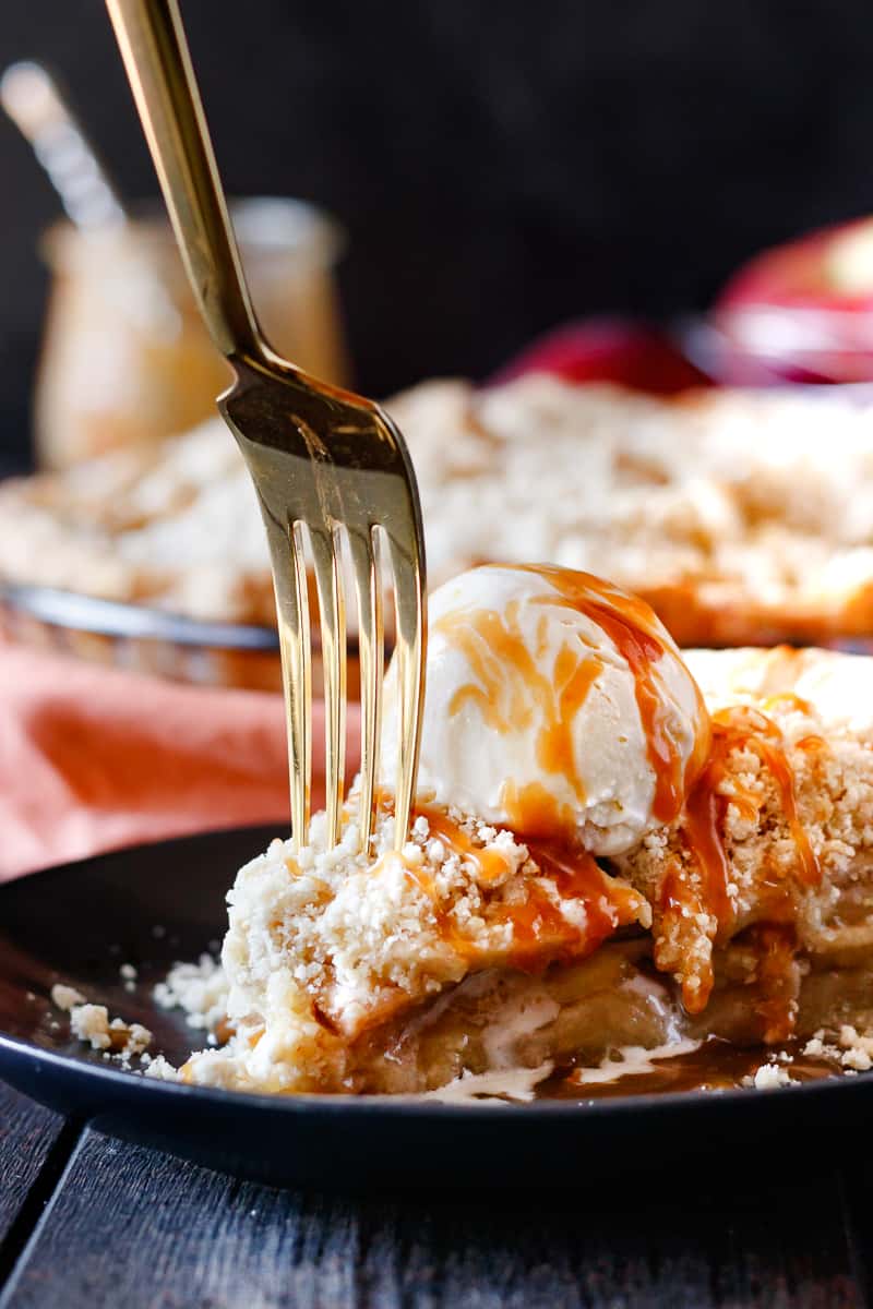 fork diving in to a slice of browned butter apple crumb pie with ice cream and caramel drips