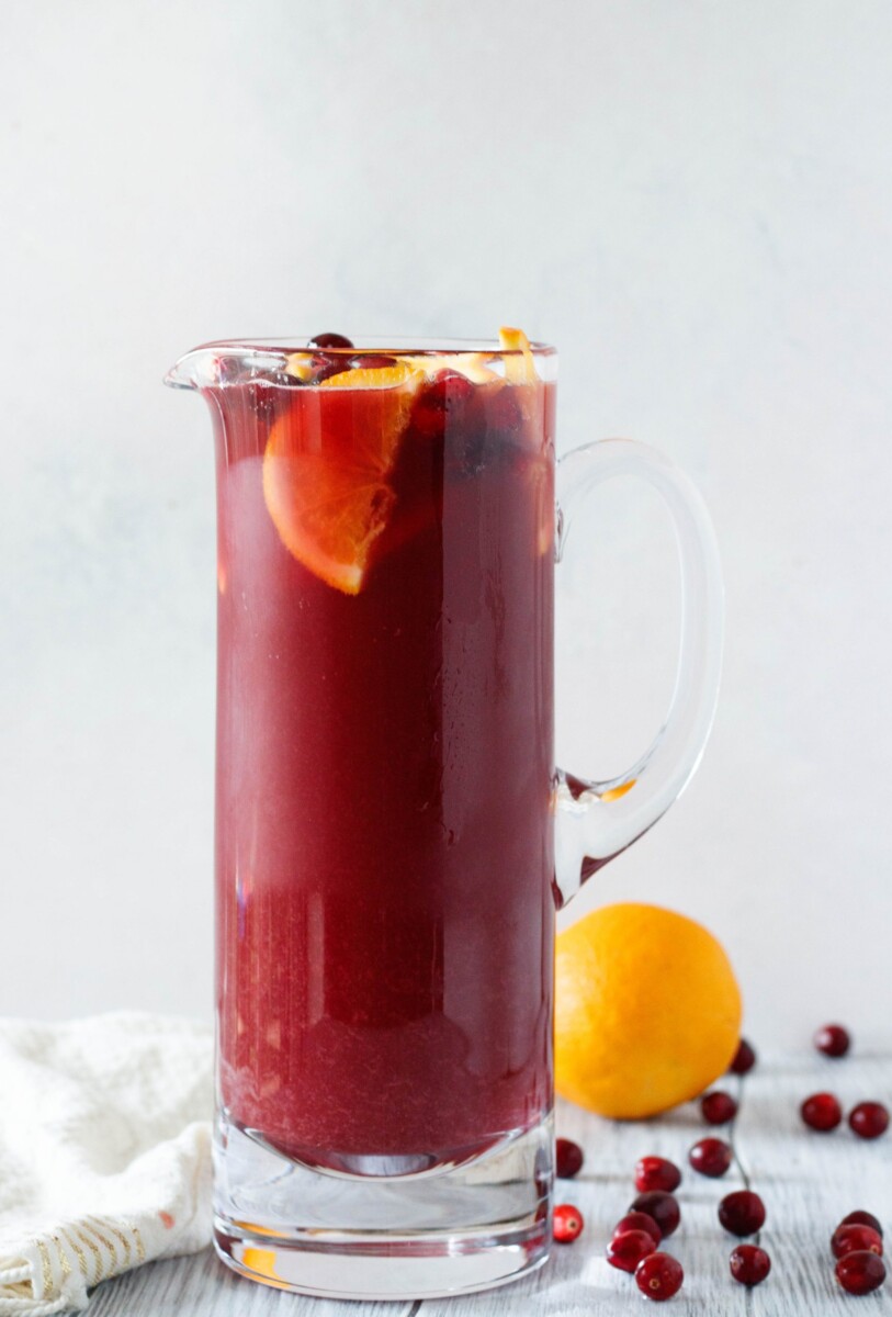 a tall pitcher of punch with an orange in the background and scattered fresh cranberries