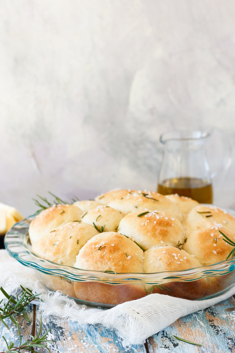 round pie dish with rosemary rolls with a light background and a small pitcher of olive oil