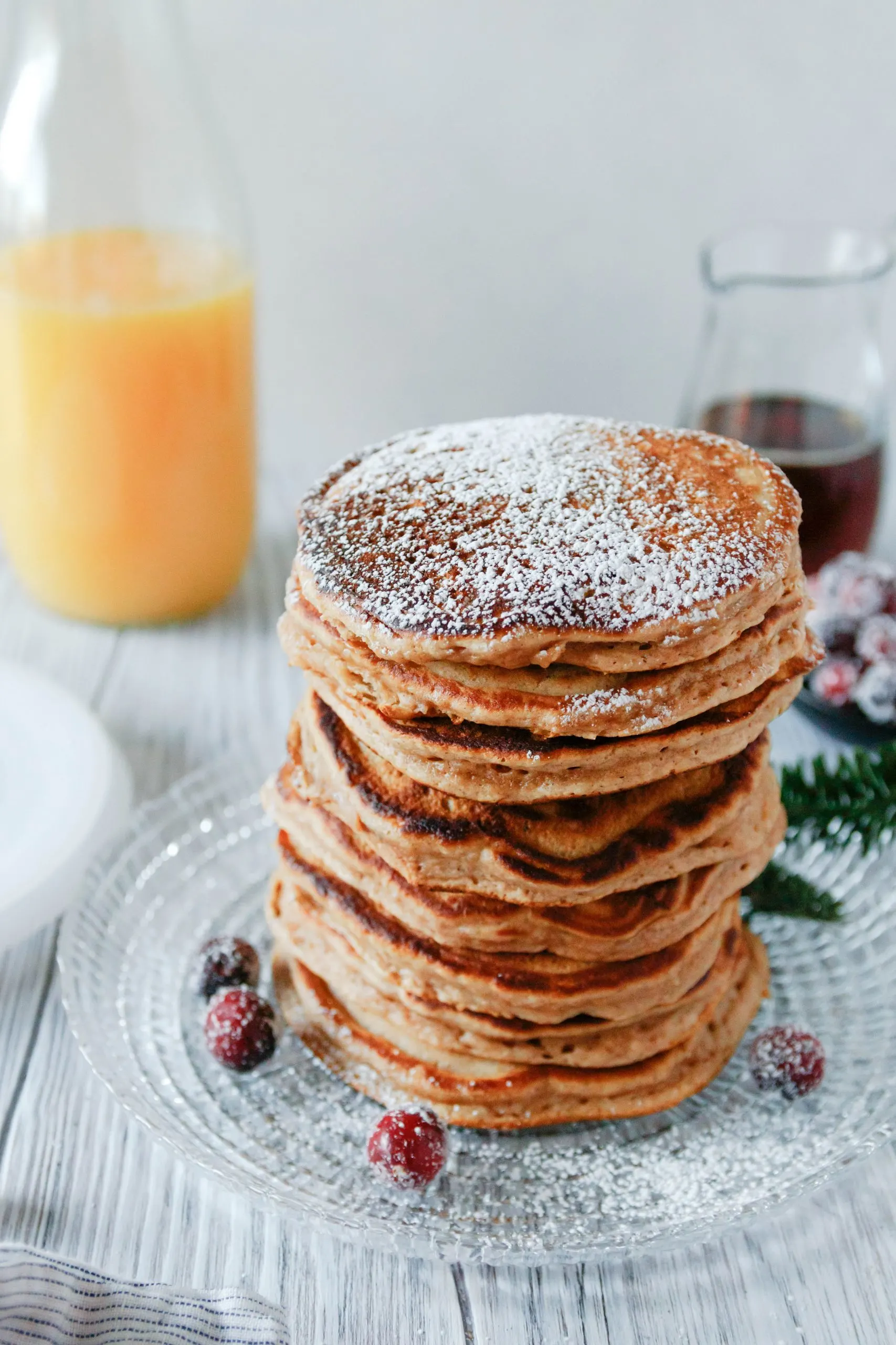 angled picture of a big stack of pancakes dusted with powdered sugar. Pancakes are on a glass plate and garnished with a few sugared cranberries.