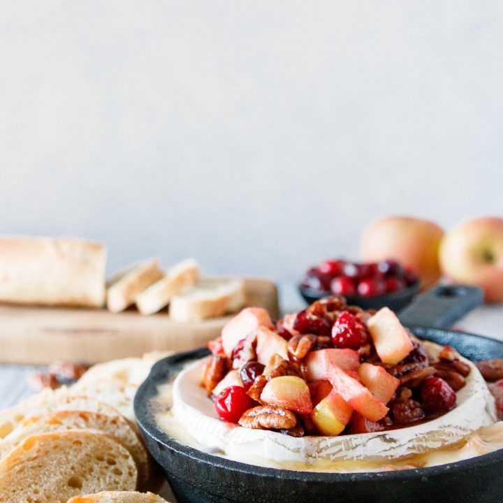 cranberry apple baked brie in a cast iron skillet with a sliced baguette and whole apples in the background