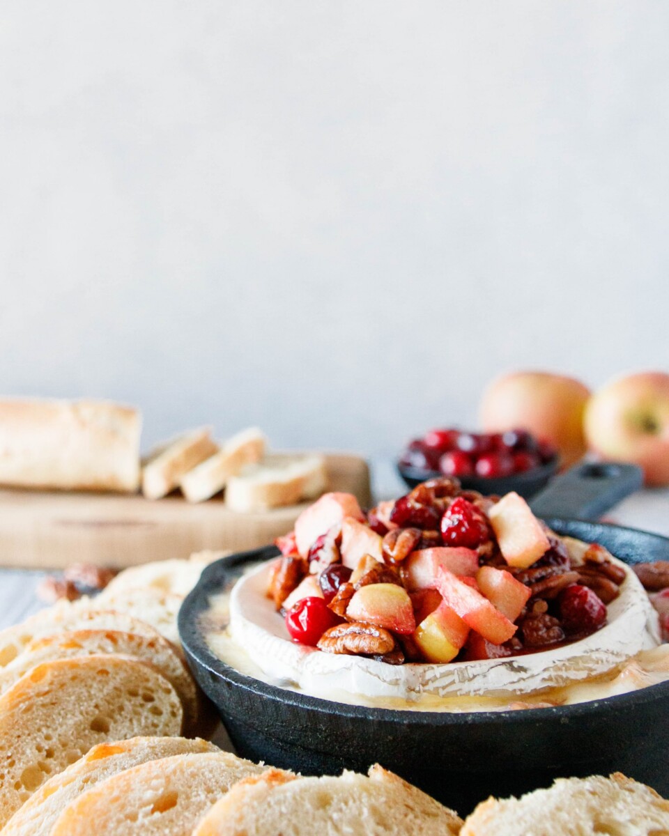 cranberry apple baked brie in a cast iron skillet with a sliced baguette and whole apples in the background