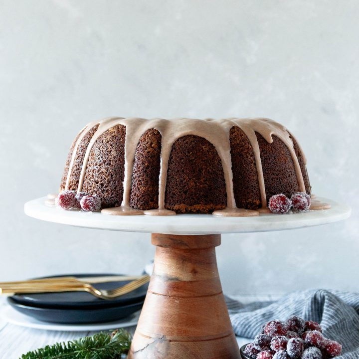 gingerbread bundt cake with cinnamon glaze on a marble and wood cake stand with sugared cranberries and greenery