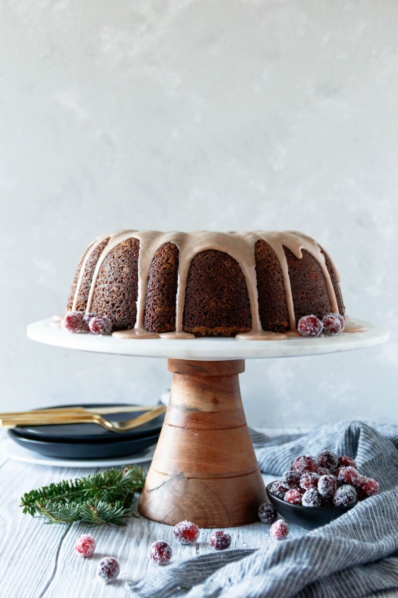 gingerbread bundt cake with cinnamon glaze on a marble and wood cake stand with sugared cranberries and greenery