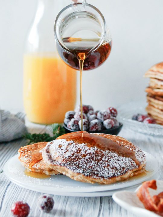 a glass pitcher pouring syrup on plated eggnog pancakes. Orange juice in the background