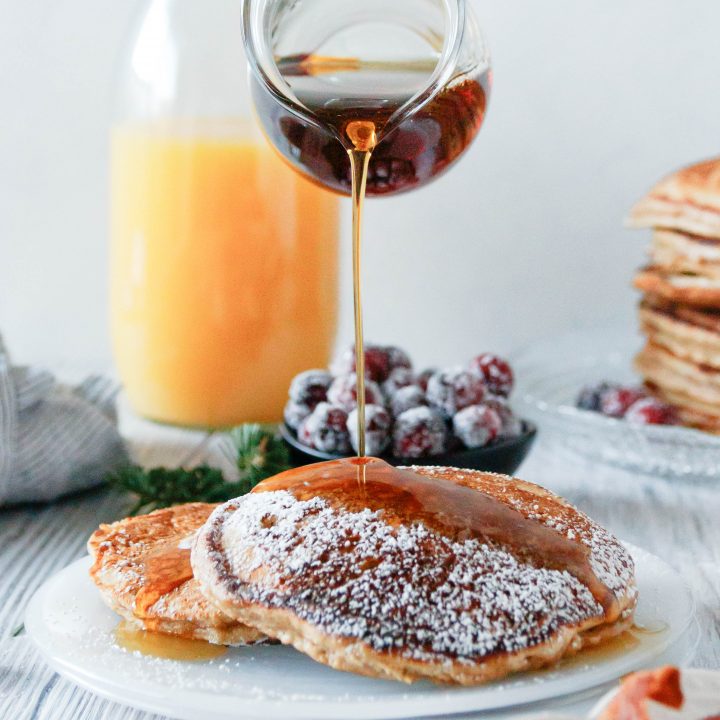 a glass pitcher pouring syrup on plated eggnog pancakes. Orange juice in the background