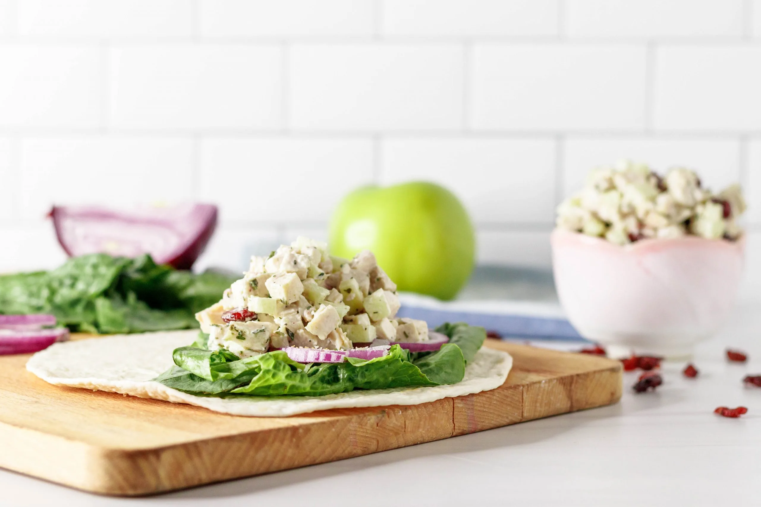 horizontal picture of the chicken salad wrap sitting on a wooden cutting board with an apple, lettuce, cut onion, and extra chicken salad in the background