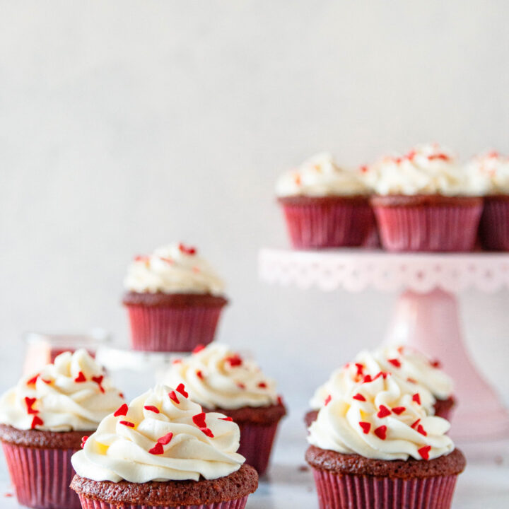 red velvet cupcakes on a marble counter with a white background. Heart sprinkles scattered on top of the cupcakes and in the work space. Some of the cupcakes are on a pink cake plate
