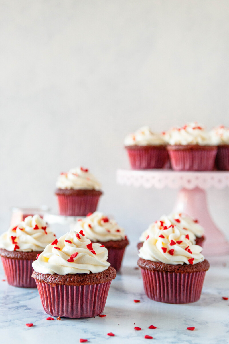 red velvet cupcakes on a marble counter with a white background. Heart sprinkles scattered on top of the cupcakes and in the work space. Some of the cupcakes are on a pink cake plate