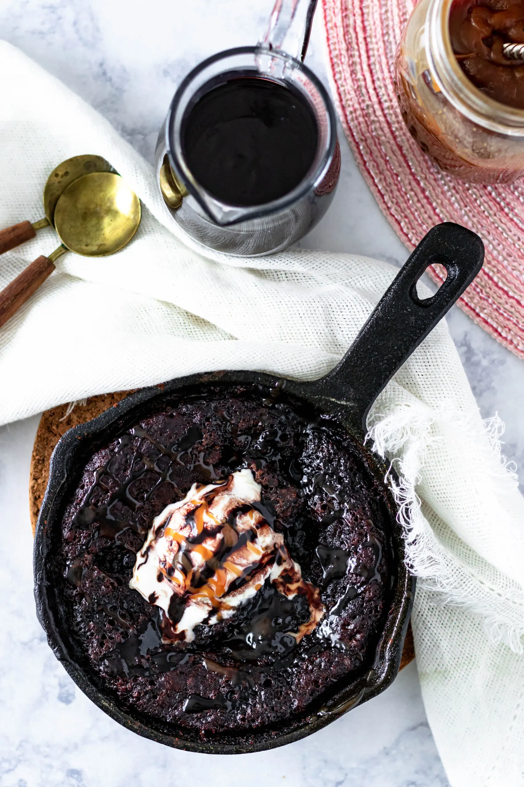 top view of the hot fudge cake accented with a napkin in the top left of the frame with gold and wooden spoons and a top view of the pitcher of chocolate syrup