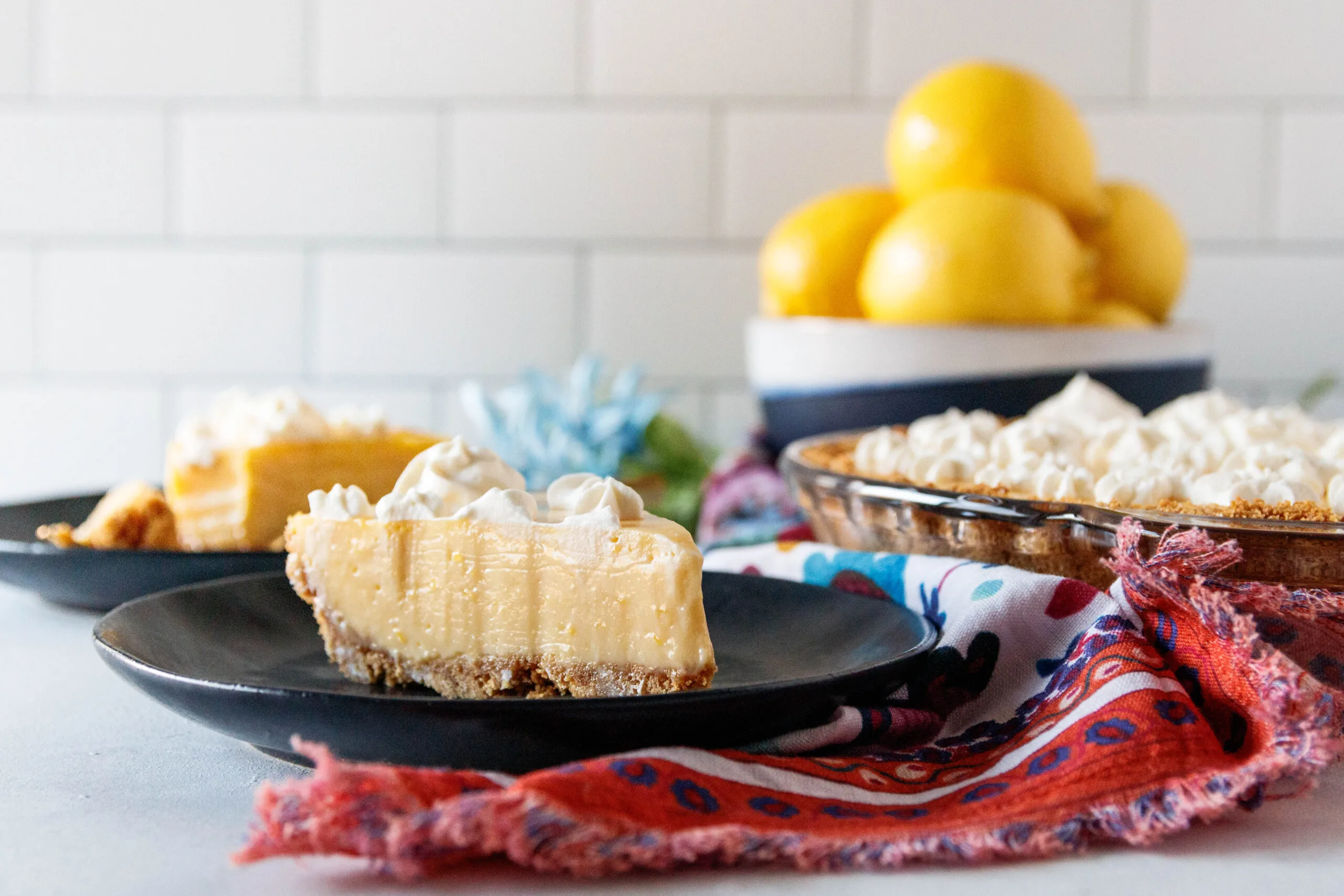 horizontal image showing a slice of lemon pie on a black plate with a bowl of lemons in the background