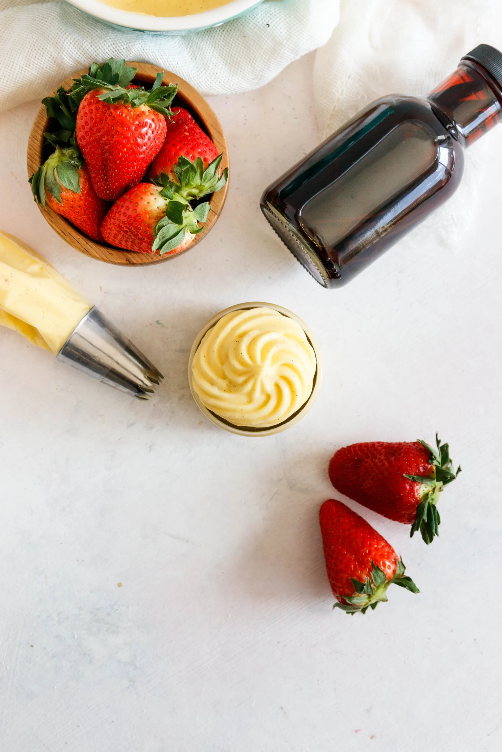 flat lay photo of the pastry cream on a textured white background. Some of the cream is in a pastry bag with a piping tip, the rest is piped into a jar. There's a bottle of extract and some scattered strawberries