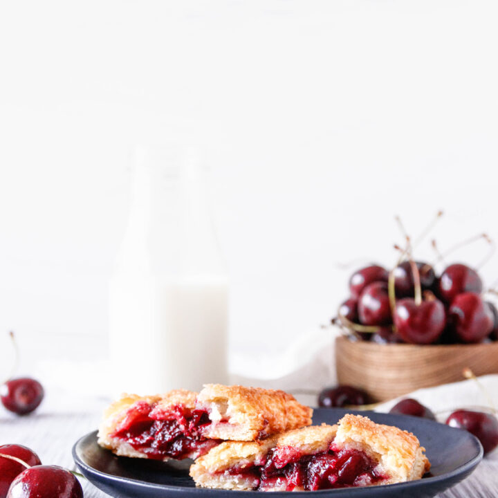 a cherry hand pie cut in half on a small black matte dessert plate. There's a bowl of fresh cherries and a small jug of milk in the background. White weathered tabletop and bright white background