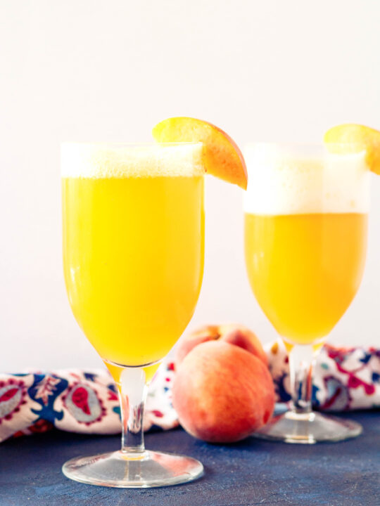 two peach shandy beer cocktails on a blue tabletop with a white background and slice of peach garnishing each glass