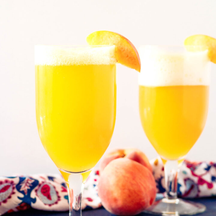 two peach shandy beer cocktails on a blue tabletop with a white background and slice of peach garnishing each glass