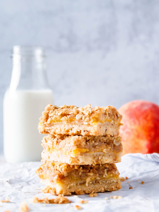 three stacked brown butter peach crumble bars on a crinkled parchment sheet. mottled light grey background