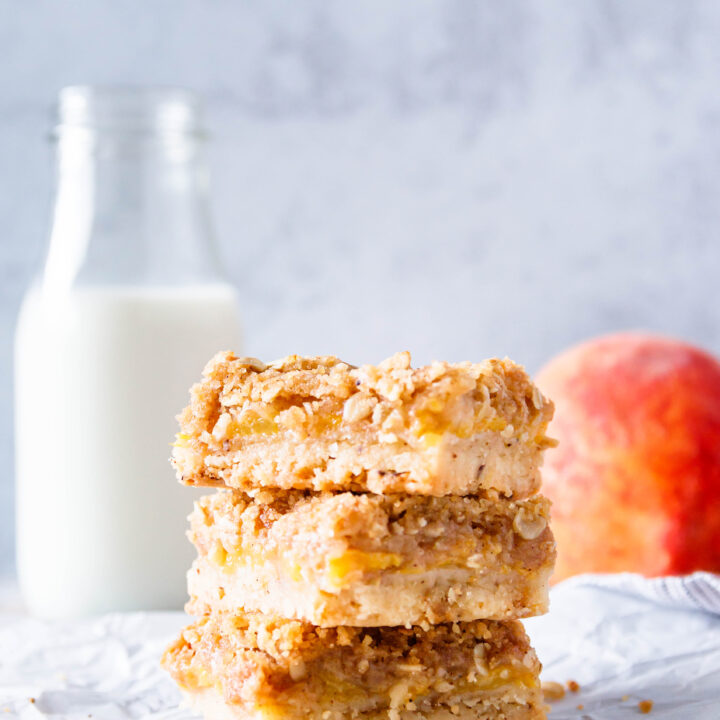 three stacked brown butter peach crumble bars on a crinkled parchment sheet. mottled light grey background