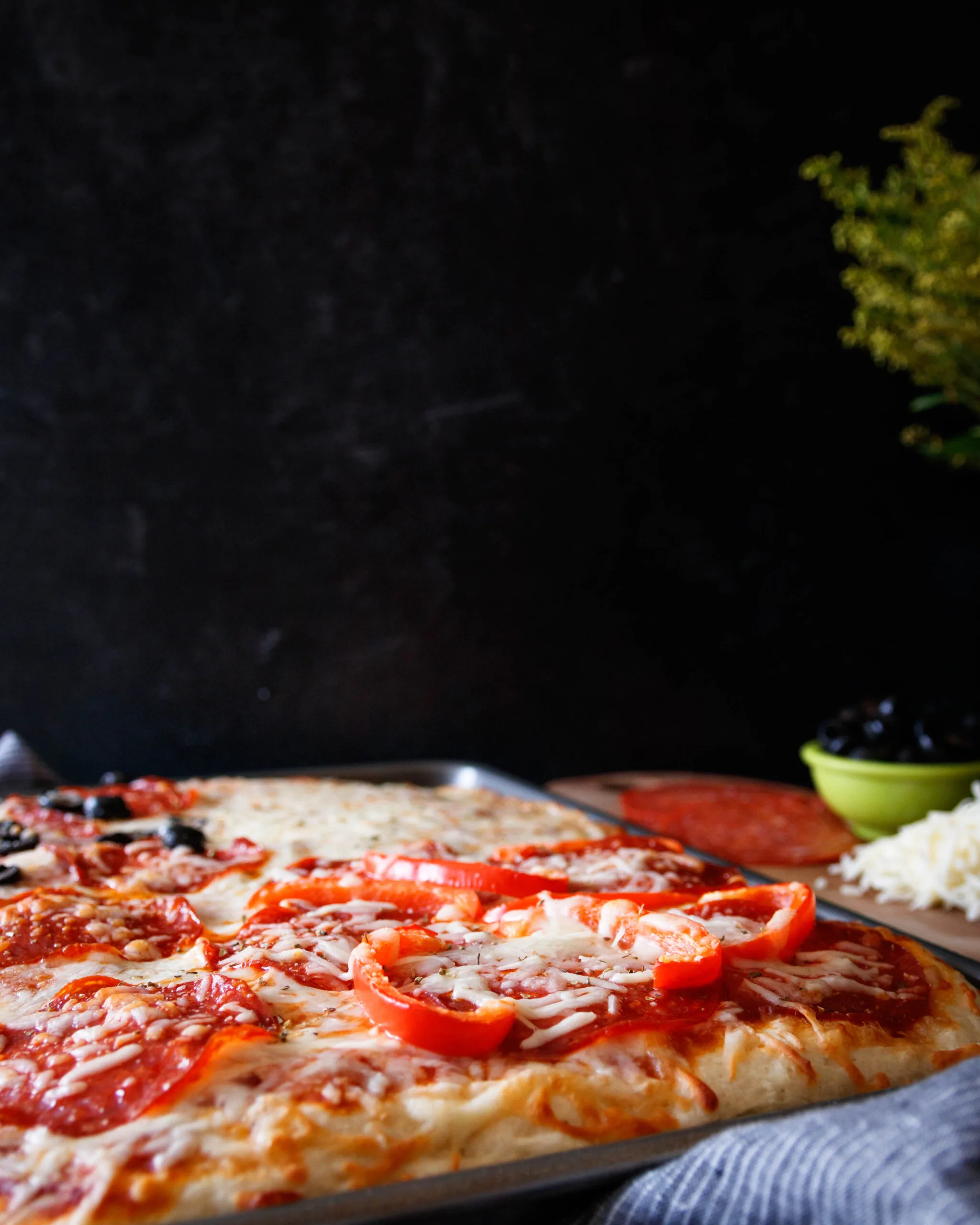 baked pizza resting in the pan on a table with olives and pepperoni in the background. Black backdrop.