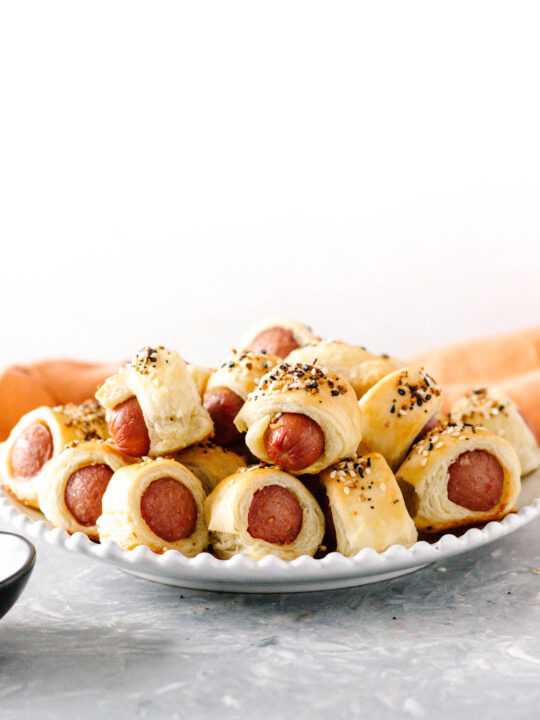 white beaded edge plate piled with everything bagel pigs in a blanket, ready to enjoy! Grey tabletop, light background, orange napkin behind the plate