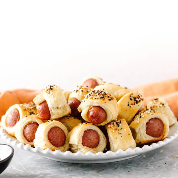 white beaded edge plate piled with everything bagel pigs in a blanket, ready to enjoy! Grey tabletop, light background, orange napkin behind the plate