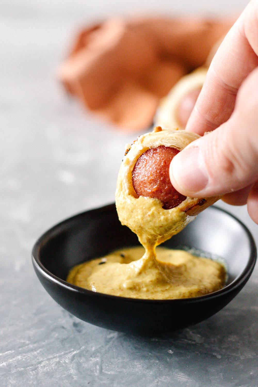 Close up of a hand dipping a mini puff pastry wrapped hot dog bite into a small bowl of mustard