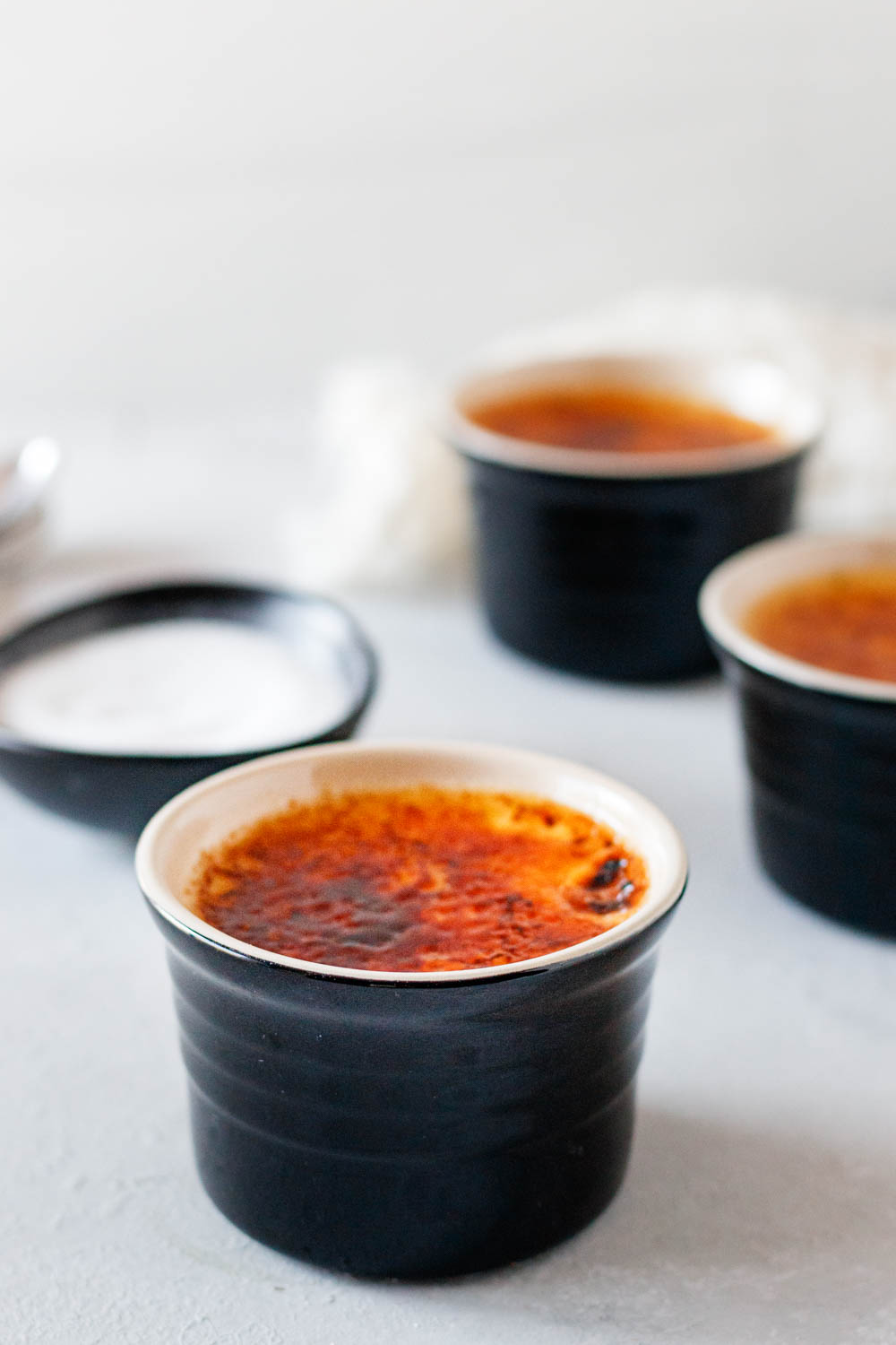 Image of 3 individual servings of Irish Cream Creme Brulee in black ramekins on a light surface with a light background. A bowl of granulated sugar for topping and creating the burnt sugar crust can be seen on the left side of the image. 