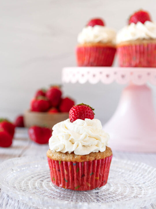 decorated strawberries and cream cupcake piled high with stabilized whipped cream frosting and topped with a small fresh strawberry. In the background there's a pink cake plate with more strawberries and a small wooden bowl of fresh strawberries
