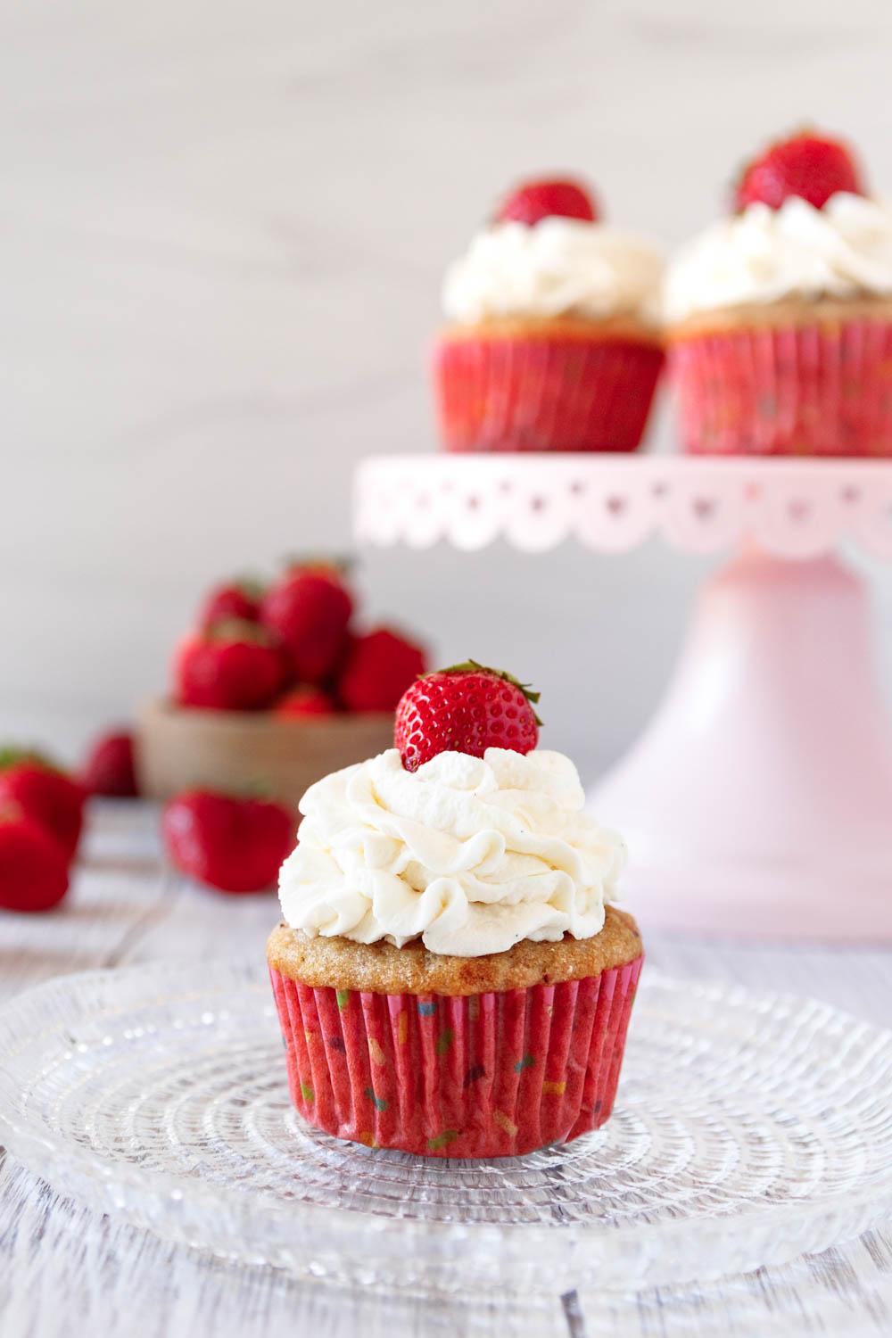 decorated strawberries and cream cupcake piled high with stabilized whipped cream frosting and topped with a small fresh strawberry. In the background there's a pink cake plate with more strawberries and a small wooden bowl of fresh strawberries