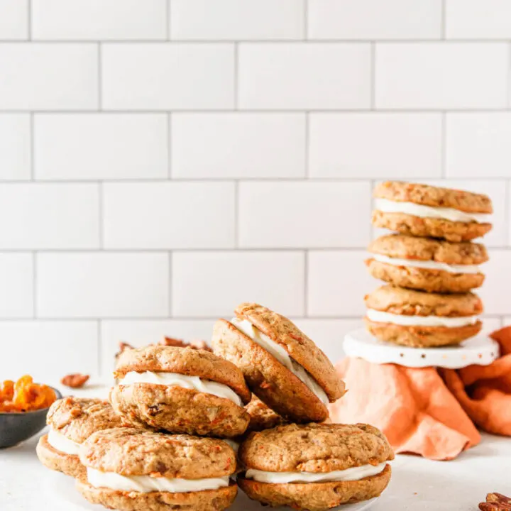 Assembled carrot cake sandwich cookies on a plate in the foreground and stacked on a white cupcake stand in the background