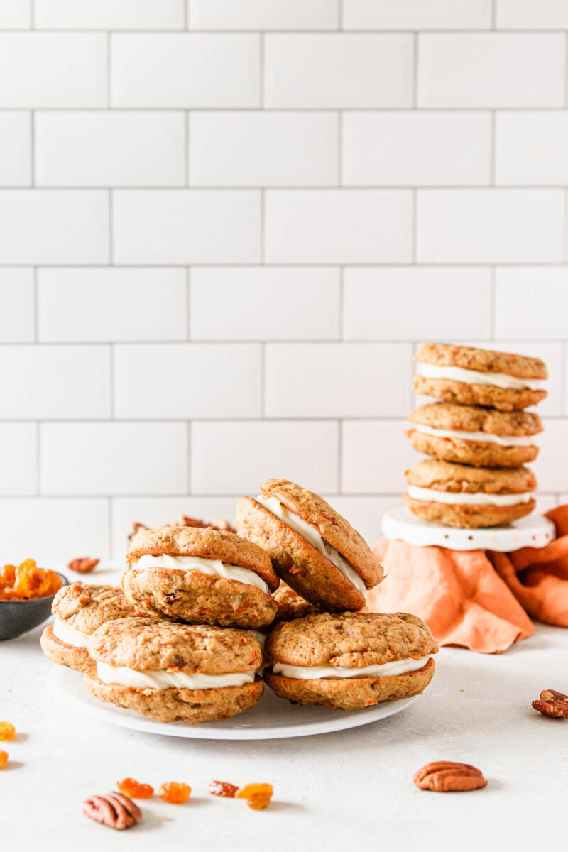 Assembled carrot cake sandwich cookies on a plate in the foreground and stacked on a white cupcake stand in the background