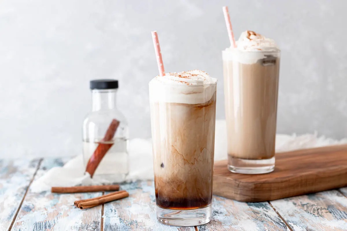 horizontal image showing 2 glasses of prepared cinnamon cold brew coffee with a whipped cream topping and pink paper straw in each cup