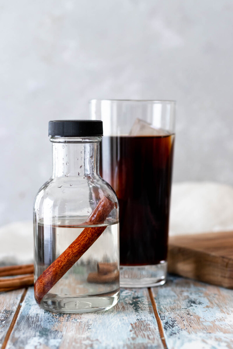 image of a small bottle of prepared cinnamon simple syrup in the foreground of the image. A black iced coffee is in the background
