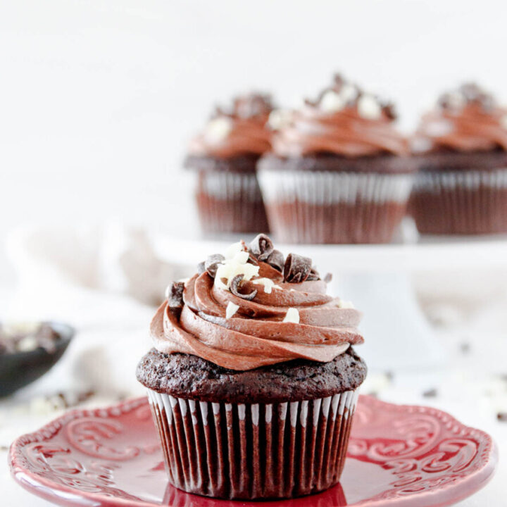 triple chocolate cupcake on a small red plate, front and center with a few more cupcakes on a white pedestal cake plate in the background