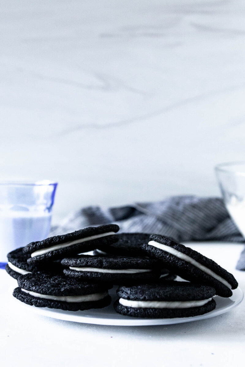 pile of homemade oreo cookies on a plate with a small glass of milk and a mixing bowl of un-piped filling in the background.